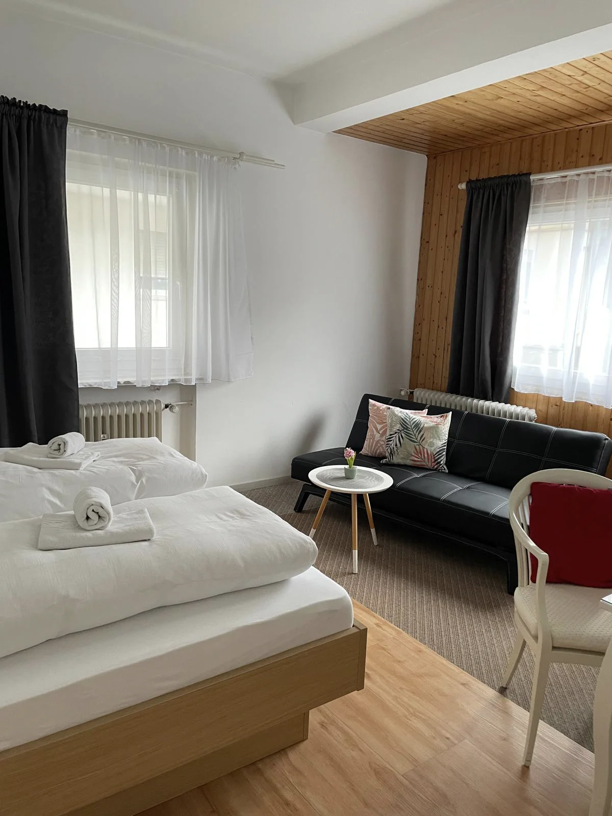 Schlafzimmer mit einem grauen Bettgestell, einem kleinem Tisch, einem dunklen Sofa und weißen Stühlen