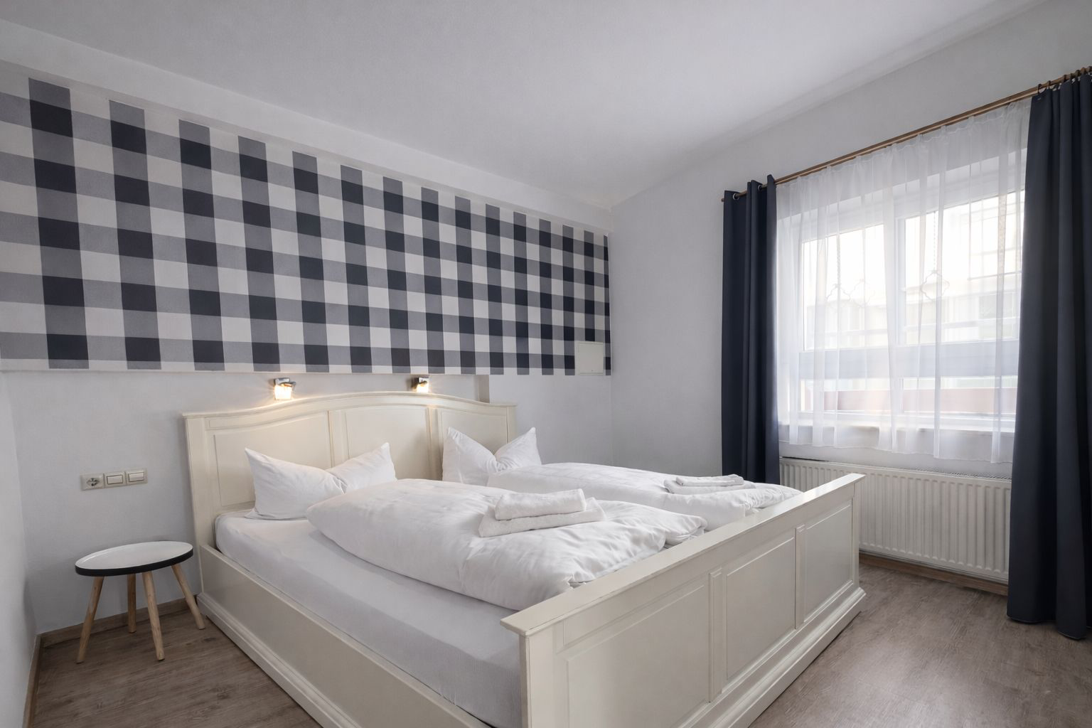 Ein Doppelbett vor einer weißen Wand mit Karo Abschnitt und dunkel blauen Vorhängen