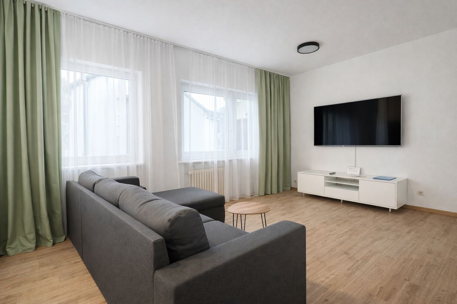 Anthrazitfarbiges Sofa, olive weiße Vorhänge und einen großen Fernseher
